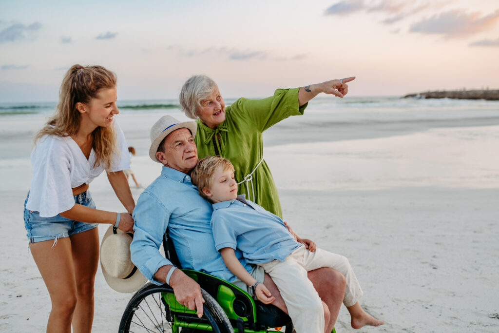 multigenerational family happy at beach. grandfather is in a wheelchair with his grandson on his lap. grandma is pointing to something off camera. mom pushing wheelchair